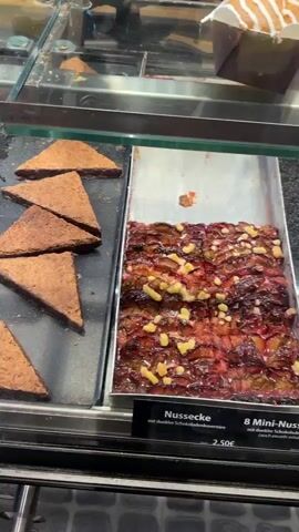 The video pans across a display case filled with various baked goods. In the foreground, several triangular chocolate pastries with a dark, crumbly topping are visible. To the right, a tray of what appears to be a nutty bar or cake with a reddish jam filling and scattered nuts is displayed. The camera then moves to show triangular slices of cake with a light brown topping, some of which are chocolate-covered. Further back, there are pastries with fruit toppings, including a strawberry-topped pastry and what look like apricot or peach Danish pastries. Other baked items, such as crumble cakes and strudels, are also present in the display. A price tag for "Nussecke" is visible. A person's legs and feet, wearing white sneakers, are visible on the left side of the frame. The environment appears to be a bakery or pastry shop.