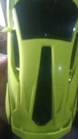 The video shows a close-up, top-down view of a toy sports car. The car is lime green with a black windshield and a black stripe down the middle of the hood. The car is held by someone's hand on the left side, and the background is blurry, suggesting it might be indoors. The camera moves slightly throughout the short clip, giving different angles of the car. The audio consists of someone exclaiming "Wow" and "mobil mainan ini bagus sekali", which translates to "toy car is very good".