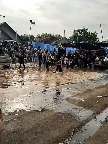 The video captures a cultural performance taking place outdoors, possibly a traditional martial arts demonstration or dance. The ground is wet and appears to be an open area with a few puddles, suggesting recent rain. A wooden pole stands prominently in the foreground, wrapped with what looks like dried banana leaves. In the background, spectators are seated on tiered bleachers, some under makeshift shelters with blue tarps. Several individuals are actively participating in the performance in the central area. One person, dressed in black and white striped attire, is in motion, possibly executing a dance or martial arts move. Another individual, in dark clothing and a head covering, stands with a whip-like object in hand, observing or participating in the performance. Banners and flags are visible in the background, adding to the festive or ceremonial atmosphere. The sky is overcast.
