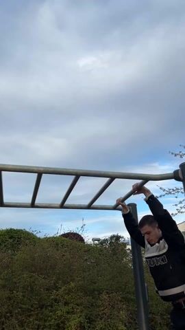 A young man is seen performing an exercise on a metal monkey bar apparatus outdoors. The man, who appears to be in his late teens or early twenties, is dressed in a black and white jacket and dark pants, with brightly colored sneakers. He is using both hands to grip the bars, pulling himself over the ladder-like structure. The sky overhead is partly cloudy, with patches of blue visible, and there are bushes and trees in the background. The camera angle is from below, looking up at the man and the sky.