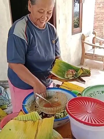 An older woman is seen in a medium shot, preparing food. She is wearing a navy blue t-shirt with grey and red accents on the collar and sleeves, and a pink skirt. Her hair is pulled back. She is standing behind a table where various food items are laid out. In her hands, she holds a banana leaf, into which she is placing a portion of food. A large metal bowl filled with noodles and a smaller bowl containing a reddish sauce are visible on the table in front of her. There are also other banana leaves scattered around. To her right, there is a white container with a red lid and a green bowl. The background suggests an outdoor setting, possibly a porch or patio, with a wall made of brick and a window. The woman is smiling as she works.
