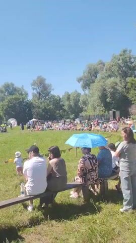 The video shows a gathering of people outdoors on a sunny day. In the foreground, several people are seated on a wooden bench, facing away from the camera. Some are wearing hats. To the left, another person in a light-colored hat is also seated. A small child in a white shirt and pink pants is standing in front of the bench.

In the middle ground, a crowd of people is spread across a grassy field. There is a long row of red and yellow flags. Further back, there is an inflatable slide with people on it and several tents. A tall pole with a flag is visible. People are standing and sitting, watching an event in the distance.

The background consists of trees and a clear blue sky. The overall scene appears to be a festival or outdoor event.