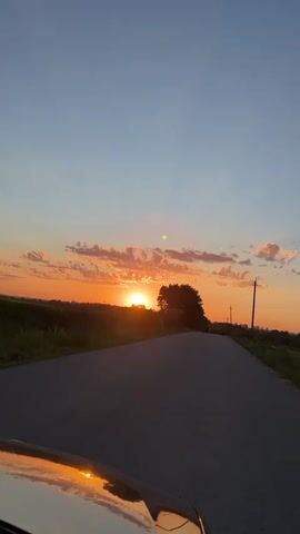 The video captures a picturesque sunset over a rural landscape in Sorotske, Ukraine. The primary subjects are a paved road curving into the distance and a line of trees silhouetted against the vibrant sky. The setting is natural and serene, with fields and vegetation visible on either side of the road.

The activity taking place is a slow drive along the road, as indicated by the perspective of the camera, which appears to be mounted on a vehicle. The front hood of the vehicle, reflecting the sunset, is visible in the lower left corner of the frame.

The time of day is clearly sunset, with the sun partially obscured by trees and casting a warm, orange glow across the sky. The clouds are illuminated by the setting sun, adding streaks of pink and orange to the otherwise clear blue sky. The weather appears to be pleasant and calm, conducive to an evening drive. There are no visible signs of people or man-made structures beyond a distant utility pole on the right side of the road. The overall emotion conveyed is one of peace, tranquility, and the beauty of nature. There is no visible text in the video.