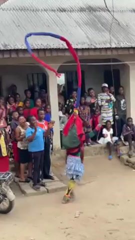 The video shows a performer dressed in a traditional costume, with a colorful headpiece consisting of a red and blue arc. The performer dances in front of a crowd gathered near a building with a gray roof and white walls.  Several people are sitting or standing on the porch. A motorcycle is parked on the left side of the frame. The ground is dirt, and there are trees in the background. The crowd appears to be enjoying the performance, some clapping and cheering. The atmosphere seems festive and cultural.