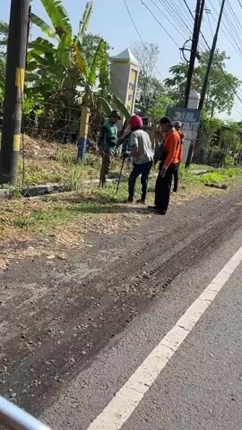 The video shows a roadside scene with several people. A man in a green shirt and dark pants is standing near a yellow and blue pole, holding what appears to be a long pole or tool. Next to him, a person wearing a red helmet and a grey shirt is holding a similar pole. Further to the right, a person in an orange shirt and black pants is standing, and another person in a dark shirt is behind them. They are on a dirt and grassy area next to a paved road. In the background, there are trees, a fence, and a sign that says "JUAL TELUR". Utility poles and wires are visible overhead. The ground on the road shows tire tracks. The lighting suggests it is daytime.