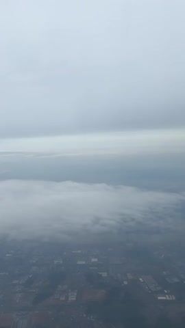 The video shows an aerial view of a city covered in fog, with a predominantly gray sky above. The city appears densely populated with buildings and infrastructure, barely visible through the thick fog layer. The upper part of the frame is filled with an overcast sky, adding to the overall grayness and obscured visibility. The shot is stable, suggesting it may have been taken from an airplane window.