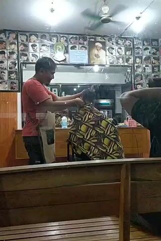 The video shows a barber cutting a client's hair in a barbershop. The barber, wearing a red shirt and an apron, is using scissors and a comb on the client, who is seated and draped with a colorful cape. The barbershop is decorated with many framed pictures of hairstyles on the walls. A ceiling fan is visible overhead. The scene appears to be filmed through a wooden partition or counter.