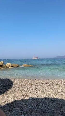 The video opens from the perspective of a pebble beach, with the foreground dominated by the texture of the stones. The clear, turquoise water laps gently at the shore, with small waves breaking. Beyond the shallow water, a larger boat with white sails and a red flag is visible in the distance. To the right of this boat, a smaller white motorboat is moving rapidly across the water, leaving a wake. In the far distance, a hazy coastline with faint hills is discernible under a clear blue sky. The overall scene depicts a bright, sunny day at a coastal location, likely a tourist destination, with recreational activities taking place on the water. There are no identifiable people in the foreground or close proximity.