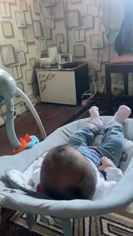 A baby, dressed in blue jeans, a white t-shirt, and pink socks, lies on its back in a grey baby rocker, with a white, orange, and blue toy near its left leg. The baby's head is towards the foreground, and its feet are towards the background, facing a large, wall-mounted television. The television displays an animated scene of a green animal playing a piano and a yellow animal playing drums. The wall behind the television and baby rocker has a grey and white geometric pattern. A small, dark table with golden accents is situated below the television, holding a white bowl and other small items. A green baby bouncer is partially visible to the left of the baby rocker.