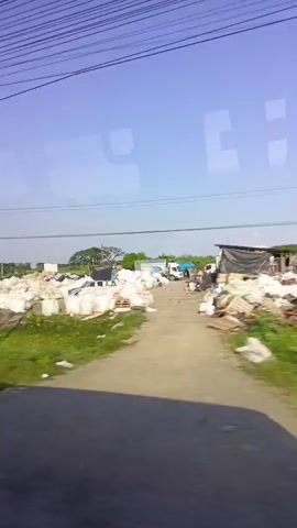 The video is taken from the inside of a moving vehicle, likely a bus or train, moving along a road. Outside the window, the landscape consists of green grassy areas and a collection of industrial or commercial establishments. These include piles of white, possibly bagged materials, trailers, trucks, and structures covered with tarps. Overhead, power lines stretch across the clear, bright blue sky. The presence of these features suggests the video was taken in an area on the outskirts of a town or city where industrial activities occur. A cellular tower is visible. A classic, possibly aqua-colored car drives down the road in the distance.