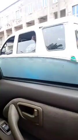 The video shows the interior of a car, with the camera looking out the side window. A white van is visible in front of the car, as well as a multi-story building with balconies. The video then cuts to a shot of the interior from the back seat, looking towards the driver and the front passenger seat. The driver is wearing a light-colored shirt and is holding the steering wheel. The front passenger appears to be wearing a light-colored shirt and has their hand raised. The car is moving, as indicated by the blurred scenery outside the windows.
