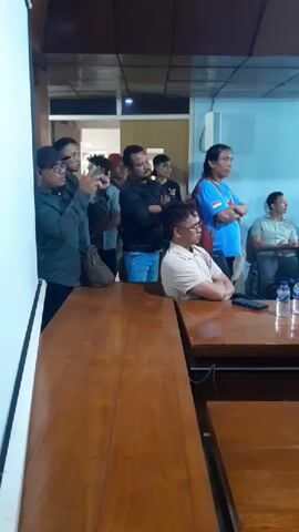 The video shows a group of men standing and sitting in a room with wooden tables. One man in the front is wearing a light-colored shirt and glasses, with his arms crossed. To his right, there are two bottles of water and a phone on a table. Behind him, a group of men are standing, some with their arms crossed. One man in the back is wearing a blue shirt with white and red accents and has his arms crossed. The room appears to be an indoor meeting or gathering space, with a light-colored screen or wall on the left side and blue walls in the background.