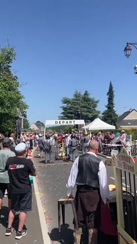 A street scene unfolds under a clear blue sky, likely a sunny day. A white banner with the word "DEPART" (which means "start" in French) is stretched across the road, indicating the beginning of an event. Crowds of people are gathered on both sides of the street, watching. Some are standing under white tents. In the foreground, a man in a white shirt, brown vest, and brown pants stands near a chair. To his right, a colorful parade float resembling a cage, labeled "CIRQUE DE RIVES," is visible. Inside the cage appears to be a stuffed animal, possibly a lion, and the word "DANGER" is displayed prominently. A person dressed as a clown, in a yellow and red checkered outfit, is standing next to the float. Confetti is scattered on the ground, suggesting the event is underway or has just begun. Several people are dressed casually, some wearing hats. The atmosphere appears festive and celebratory.