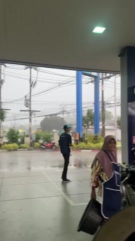 The video opens with a wide shot of a gas station with a covered canopy. It is raining heavily, and the ground is wet. A man wearing a blue shirt and black pants walks across the frame from left to right, carrying a backpack. To the right, a woman in a pink headscarf and apron stands near a gas pump. Several motorcycles are parked under the canopy. In the background, traffic can be seen on a wet road, and the sky is overcast. The camera pans to the right, showing more of the gas pumps and a few more people on motorcycles. The video continues to pan, revealing more of the surroundings, including houses and palm trees through the rain. The scene is bustling with activity despite the inclement weather, with people coming and going from the gas station.
