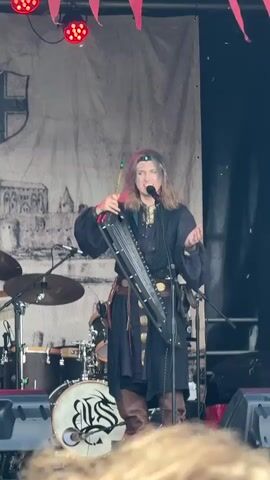 A person with long blonde hair and face paint stands on a stage playing a nyckelharpa and singing into a microphone. They are wearing a black medieval-style tunic, brown leather boots, and a silver headband with a jewel. Behind them is a drum set with a logo on the bass drum and a large banner depicting a castle. Red stage lights are visible above the banner. The person is holding a bow in their right hand, which they are using to play the instrument. Their left hand is on the strings and keys of the nyckelharpa. They are looking towards the audience, with their mouth open as if singing. The stage is set up outdoors, with some lighting equipment visible in the foreground. The overall scene suggests a musical performance at an outdoor festival or event.