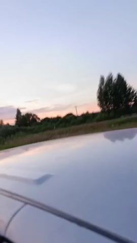 A first-person perspective video shows the view from the roof of a car driving on a road at sunset. The sky is a mix of blue, pink, and orange hues. There are clouds in the distance. Trees and fields line the side of the road. A road sign is visible at one point. The car's roof is in the foreground, reflecting the sky and surroundings. The video appears to be taken during a leisurely drive through the countryside.
