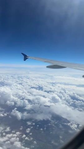 The video depicts a view from an airplane window, showcasing the aircraft's wing extending into a clear blue sky. Below, a dense layer of white, fluffy clouds stretches out, partially obscuring the ground below. Patches of green landscape can be seen through breaks in the cloud cover. The camera appears stable, suggesting a smooth flight.