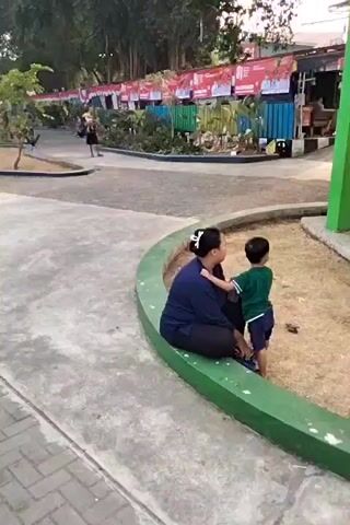 A woman and a child are sitting in a park. The woman is wearing a blue shirt and dark pants. The child is wearing a green shirt and blue shorts. The woman has her arm around the child. There are trees in the background and a fence with banners. The ground is covered in concrete and sand.