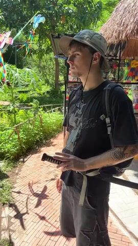 A young man wearing a black t-shirt, grey shorts, a bucket hat, and a backpack stands in a sandy area with red brick paving. He is holding a long, thin, white object that resembles a sword. He moves the object in a circular motion in front of him and then raises it above his head while extending his left arm. In the background, there are trees, plants, and some colorful kites hanging from a line. A thatched-roof structure with shelves displaying merchandise is visible to the right. Shadows of the kites are cast on the ground.