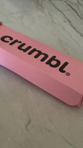 Here is a description of the video:
A pink Crumbl cookie box is opened on a gray and white patterned surface. The box contains four large cookies. The first cookie is frosted with white frosting and covered in colorful sprinkles. The second cookie is frosted with light brown frosting, drizzled with dark chocolate, and topped with nuts. The third cookie is a blueberry cookie with a crumble topping.