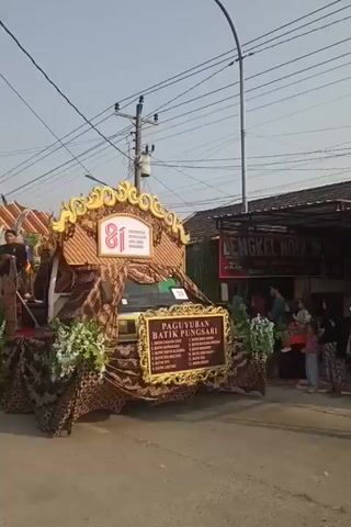 A street parade float is decorated with batik fabric and adorned with flowers and a large sign. The float is on a street, and people are watching the parade. The sky is clear and the sun is shining. A sign on the float reads "PAGUYUBAN BATIK PUNGGARI."