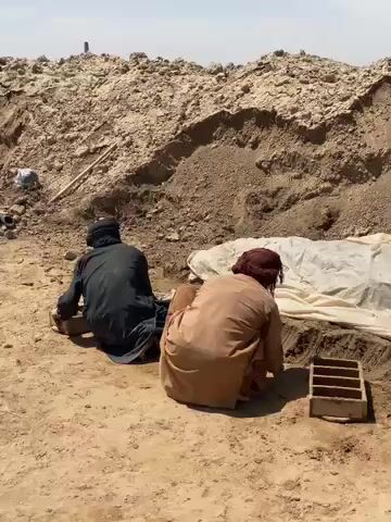 Two men are depicted working outdoors in a dry, dusty environment, likely in Deh-E Bagh, Afghanistan. The scene features a large, excavated pile of light brown earth in the background, suggesting a construction or excavation site. The ground is sandy and barren.

One man, wearing a brown traditional outfit and a maroon turban, is squatted on the right side of the frame. He is actively engaged in pressing material, likely mud or earth, into a rectangular wooden mold on the ground. A second, identical wooden mold is visible to his right, already filled.

The second man, dressed in a black traditional outfit with a black head covering, is squatted on the left. He is also positioned near a wooden mold, though his exact action is less clear in this moment. Both men appear focused on their task.

The activity suggests the manual creation of mud bricks or similar earthen building materials. The bright, clear sky indicates daytime, likely sunny weather, in what appears to be an arid or semi-arid region. No visible text is present in the scene. The overall atmosphere is one of manual labor in a desolate, sun-drenched landscape.