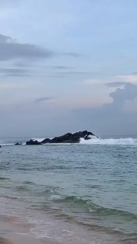 The scene depicts a beach with gentle waves lapping the shore. In the distance, a rocky islet stands in the ocean, with waves crashing against it. The sky is overcast with soft, diffused light. Two individuals, who appear to be children or teenagers, run along the shoreline, moving from right to left in the frame. A palm tree is visible on the left side of the frame.