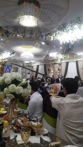 The video shows a dimly lit indoor event space that appears to be a banquet hall or reception venue. The ceiling is adorned with ornate chandeliers and decorative floral arrangements. Several tables are set with food and drinks, and many people are seated, suggesting a gathering such as a wedding reception or formal dinner. The attendees are dressed in formal attire, including men in suits and women in dresses. Some individuals are standing and interacting, while others are seated and observing. The overall atmosphere seems festive and celebratory.