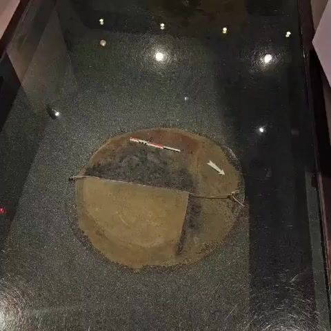 The video shows an overhead view of a circular archaeological artifact, possibly a shield or a large ceramic object, displayed under glass. The artifact is partially divided into two sections, one darker and appearing more weathered, the other lighter and smoother. A measuring scale with red and white markings is placed across the darker section, and a white arrow points towards the right side of the artifact. A thin rope is attached to the artifact and extends to the right edge of the frame. The artifact is set against a textured dark surface, possibly a floor or a display base. Lights are reflected on the glass surface, indicating the display environment.