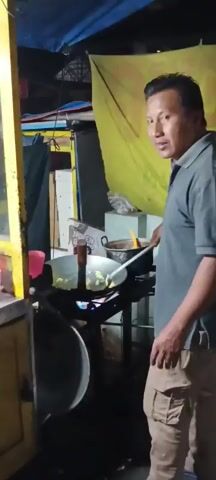 A man is cooking at a food stall. He is wearing a grey shirt and khaki shorts. He is using a wok over an open flame. In front of him, there are containers of chopped vegetables, including lettuce, tomatoes, and cucumbers. There are also plates and bowls. Behind him, a yellow tarp is visible. The man raises his hand to block the camera. A sign behind him reads "NASI GORENG & MIE CAPCAY."