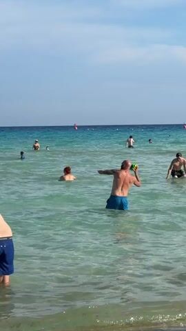 The video captures a bright, sunny day at a beach, likely in Ayia Napa, Cyprus, given the context. The scene features multiple people enjoying the clear, turquoise water of the sea.

In the initial moments, a bald man wearing blue swim trunks is seen playing with a colorful beach ball in the shallow water, with his arms outstretched as if to catch or throw it. Other individuals are visible in the background, some swimming or standing further out in the water. The sky is a clear blue with minimal clouds.

As the camera pans slightly, a group of young men can be seen standing and interacting in the water further offshore. Closer to the foreground, another man, seen from behind, is bending over in the shallow water, wearing blue "JACK & JONES" brand swim trunks. The colorful ball continues to float in the water.

Towards the end of the video, a woman in a patterned bikini walks out of the water towards the shore, with the floating ball near her. The water throughout the video remains calm with small ripples and is exceptionally clear, revealing the seabed. The overall atmosphere suggests a relaxed and enjoyable day at the beach.
