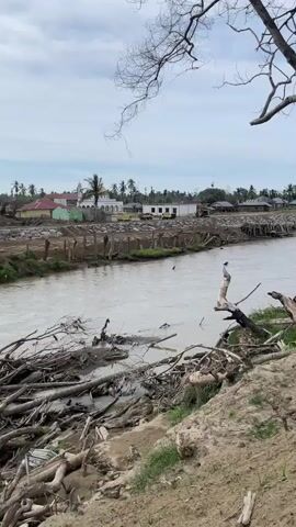 A muddy river flows through a scene of destruction and rebuilding. The river is filled with a large amount of debris, including branches and logs. On the banks of the river, there is more debris and muddy ground. In the background, a village appears to be under construction or repair. There are several buildings visible, some with colorful roofs, and a mosque with a white dome. Some of the buildings appear to be damaged, with exposed foundations and incomplete structures. There is evidence of heavy machinery, such as a bulldozer, in the background, suggesting ongoing work. The sky is overcast with clouds.