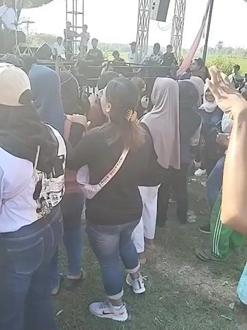 Here is a description of the video:

The video shows a crowd of people watching a live band performing on a large outdoor stage. The stage is covered and has several speakers and lighting rigs. The band consists of multiple members, including a female singer in a black dress. Many people in the crowd are filming the performance with their phones. The video appears to be taken from within the crowd, with the camera occasionally moving to capture different angles of the stage and the audience. The setting seems to be an open area, possibly a field, with greenery visible in the background. The weather appears sunny.