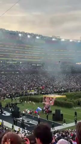 The video shows a large crowd gathered in a stadium, likely for a major sporting event. A performance is underway on the field, with dancers and flags visible. The stadium is packed with spectators, and there is smoke or haze in the air. A large screen displays the name "PERRE" above the Levi's Stadium sign. People in the foreground appear to be recording the event with their phones.