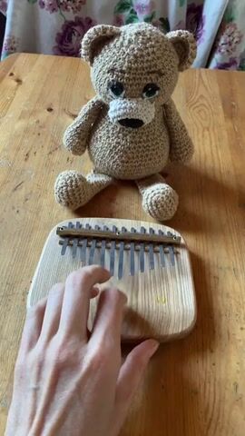 The video shows a close-up shot of a person's hands playing a kalimba. The kalimba is made of wood with metal tines. The person's fingers are shown plucking the tines to produce music. In the background, a crocheted teddy bear sits on a wooden table. The table is set in front of a floral curtain. The lighting is natural and appears to be from a window. The video is silent except for the music from the kalimba.