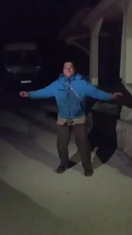 A man stands outside at night, his arms outstretched. He wears a blue jacket, brown pants, and dark shoes. A white van is parked behind him, and a building with a porch is to his right. The man's shadow is cast on the ground in front of him. The camera then points upward, showing an indeterminate object. Music is playing.