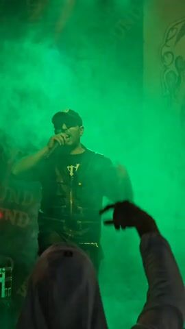 A man in a black t-shirt, vest, shorts and baseball cap is rapping on stage in front of a green screen.  He is holding a microphone in his right hand and gesturing with his left. A fan’s raised arm and finger are visible to the right of the stage, as well as a person in the foreground, wearing what seems to be a white hoodie. Behind the rapper are the words “Underground Sound” flashing in red lights. When the camera zooms out, a logo becomes visible behind the rapper that says “The Riders MC Bulgaria.” There are green lights shining from above the stage.