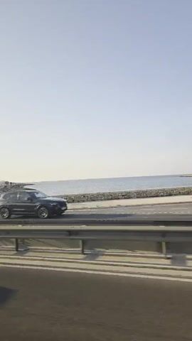 From the interior of a moving vehicle, the camera records a coastal highway. The highway runs alongside a stone barrier with the sea and the shoreline visible beyond. The sky is clear and bright. Vehicles including a dark green SUV and a silver hatchback pass on the adjacent lane. People are visible beyond the stone barrier, including a person jogging shirtless.  A light pole is prominently featured in the foreground.