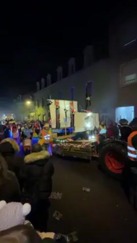 The video shows a nighttime parade in a town. People in warm coats are watching a float. The float is pulled by a red tractor and decorated with lights. People are dressed in costumes in the parade and walk along the street. The buildings on the street are lit up. The weather appears to be cold. The video is taken at eye level from the crowd.