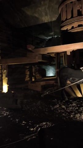 The video shows a dimly lit interior of what appears to be a mine. The focus is on a large wooden wheel with rope wrapped around it. To the right of the wheel is a wooden structure that seems to be part of a mechanism involving a mannequin of a horse. The horse mannequin is black and wearing what appears to be a harness. The walls are rough and made of rock. The scene has a historical or industrial feel.
