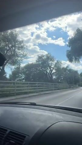 The video is taken from the inside of a car driving on a road on a sunny day with puffy white clouds in a bright blue sky.  There are trees lining the road, separated from the pavement by a white metal guardrail.  Another car is driving in front of the camera car.  The dashboard is visible with air vents and a speedometer.  The interior of the car is dark.