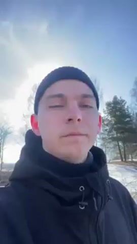 The video shows a person wearing a black beanie and a black hooded jacket standing in a snowy outdoor environment. The sun is shining brightly, creating a glare. The ground is covered with snow, and there are trees and some grass visible in the background. The person is looking at the camera. The camera pans slightly to the right, showing more of the snowy field and a paved path.