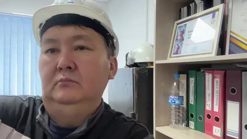 A medium shot shows a man wearing a white hard hat and a dark vest or jacket. He is looking directly at the camera with a neutral expression. The background is a bookshelf filled with binders and framed documents. A bottle of water is also visible on the shelf. The lighting suggests an indoor environment, possibly an office or a workshop. The man's facial features and the contents of the bookshelf indicate a formal or professional setting.