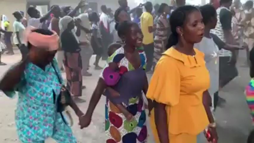 A crowd of people dance in a dusty clearing. Most people are dark-skinned. The woman at the right wears a yellow top. In the center, a woman in a colorful dress carries a child in a purple wrap. Another woman, also in a colorful dress, stands to the left. There are buildings in the background. The mood seems celebratory.