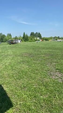 The video shows a grassy airfield on a sunny day. There are several light airplanes parked on the grass. A person is walking across the field towards the planes. There are some trees and buildings in the background. The camera pans to show more of the parked airplanes. The sky is blue with some scattered clouds.