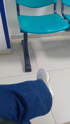 The video shows a row of light-blue, plastic chairs fixed to a white wall with a blue stripe. The chairs appear to be in a waiting area. A person wearing white shoes and blue pants walks by. The camera angle is low, suggesting it is being held near the ground. The lighting is bright, and the scene appears clean.