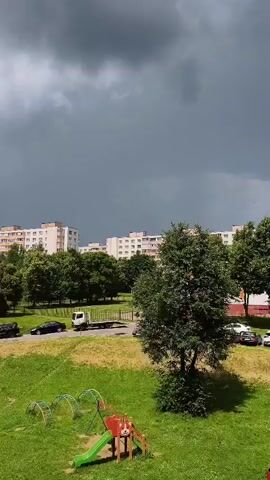 The video shows an outdoor scene under a dark, cloudy sky. In the foreground, there is a grassy slope with a children's playground structure that includes a slide. Further back, there is a paved road with several cars parked and driving. Beyond the road, a row of trees separates the road from apartment buildings. The apartment buildings appear to be multi-story residential structures. The sky is filled with heavy, dark clouds, suggesting an impending storm. The overall lighting is dim.