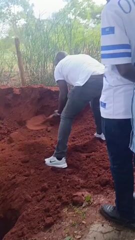 The video shows a group of people gathered around a freshly dug rectangular grave in a yard with red soil and some greenery. A man wearing a white shirt, gray pants, and white sneakers is using a shovel to put soil back into the grave. Other people are standing around the edges of the grave. Some are wearing white shirts with blue accents. The overall scene suggests a somber occasion, likely a burial.