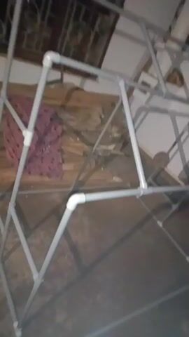 The video begins with a close-up of a PVC pipe structure, then pans to a hand touching one of the pipes. The scene then shifts to a blurry shot of a person wearing an orange shirt, followed by a dimly lit area with what appears to be construction materials and insulation. Finally, the video shows a young man in a white t-shirt looking at his phone in an outdoor setting with a metal roof overhead.