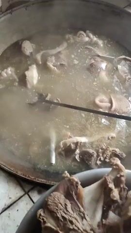 The video shows a close-up view of a large pot filled with a bubbling broth and various pieces of meat and what appear to be animal organs. A metal utensil with a forked end is being used to stir or move the contents of the pot. In the foreground, a white bowl filled with cooked meat pieces, including bones and gristle, is visible. The scene appears to be in a kitchen or food preparation area, with a tiled surface visible at the bottom left. The lighting is somewhat dim, and steam rises from the pot, indicating that the food is hot and cooking. The focus is on the cooking process and the ingredients in the pot and the bowl.